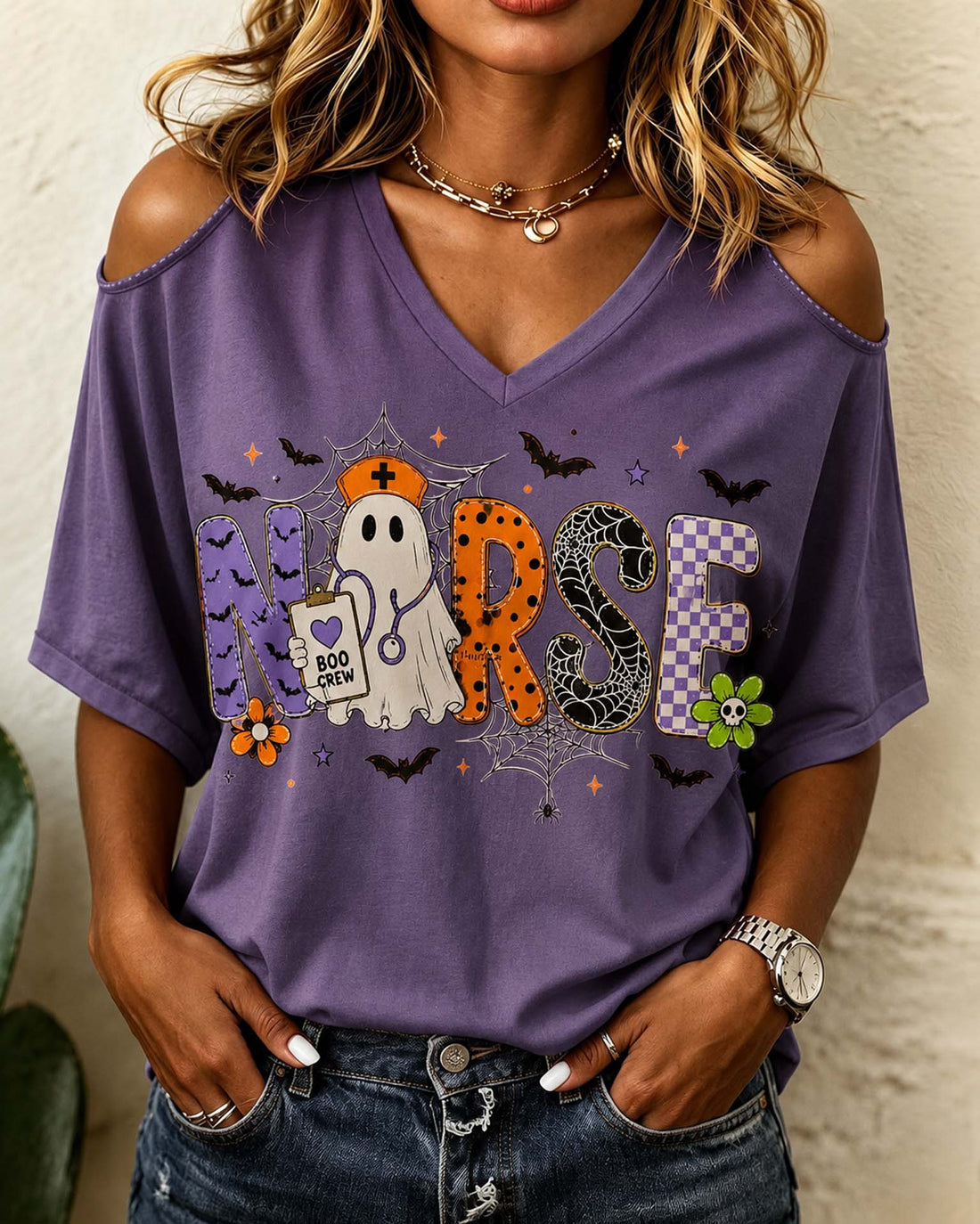A woman wearing a purple cold-shoulder V-neck t-shirt with a Halloween-themed 'Nurse' graphic, paired with blue jeans.