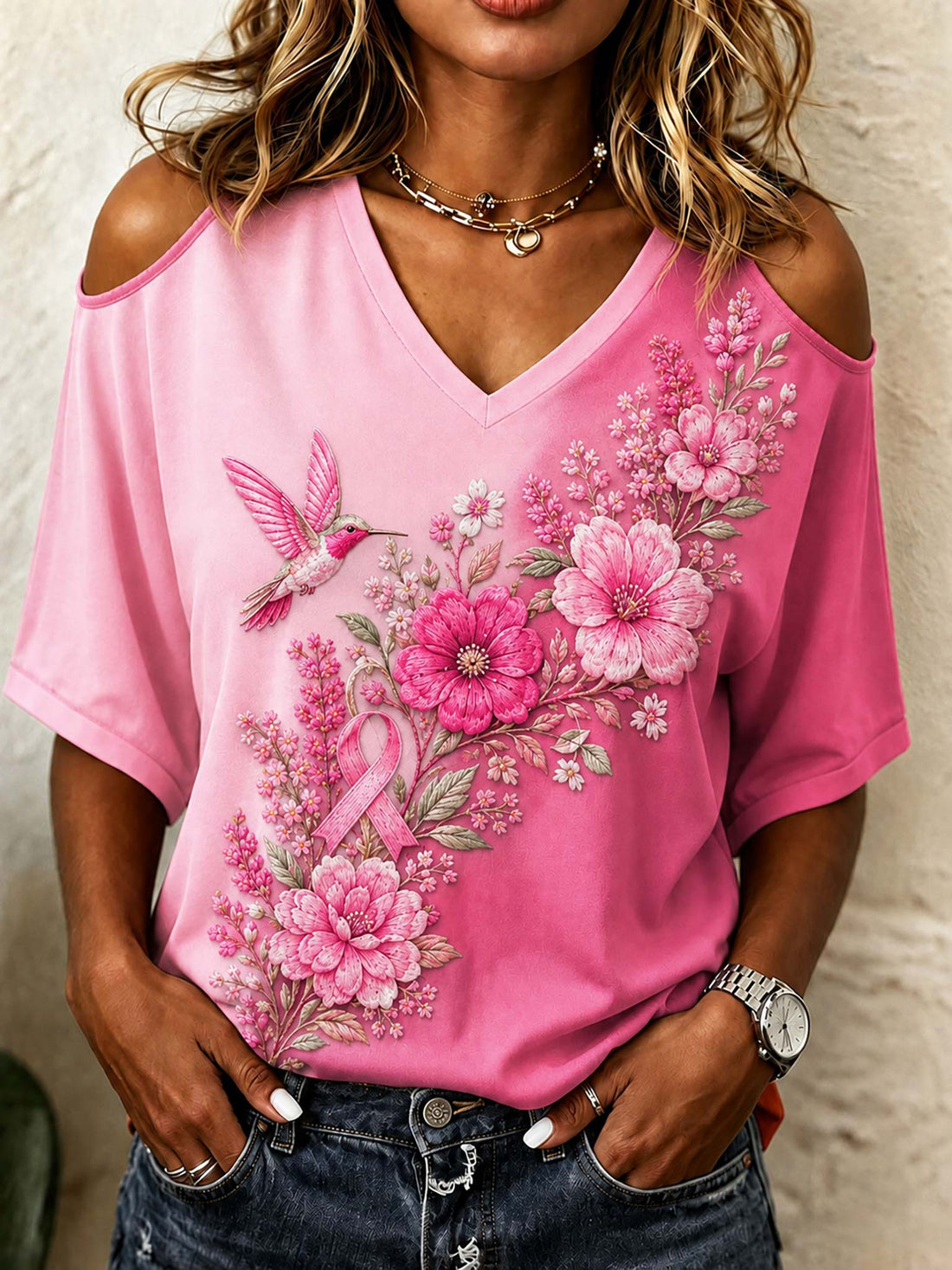 A woman wearing a pink cold-shoulder V-neck t-shirt with a large pink floral and hummingbird graphic on the front, paired with dark blue jeans.