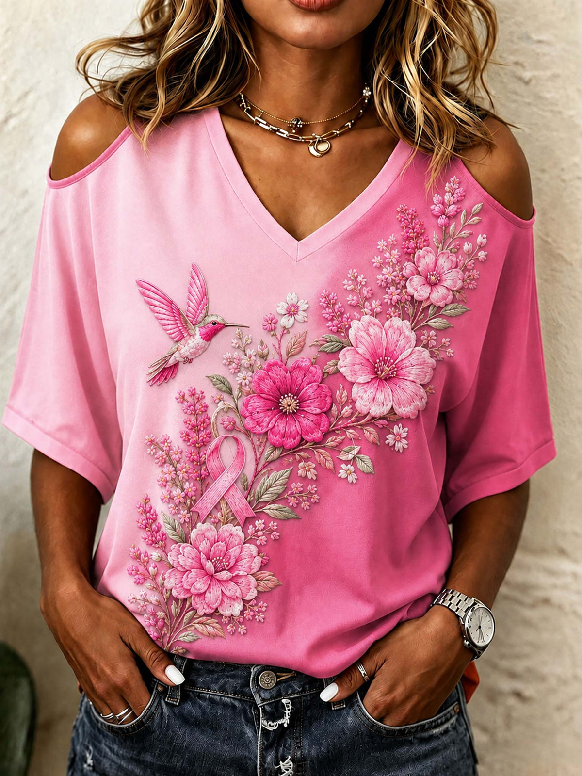 A woman wearing a pink cold-shoulder V-neck t-shirt with a large pink floral and hummingbird graphic on the front, paired with dark blue jeans.