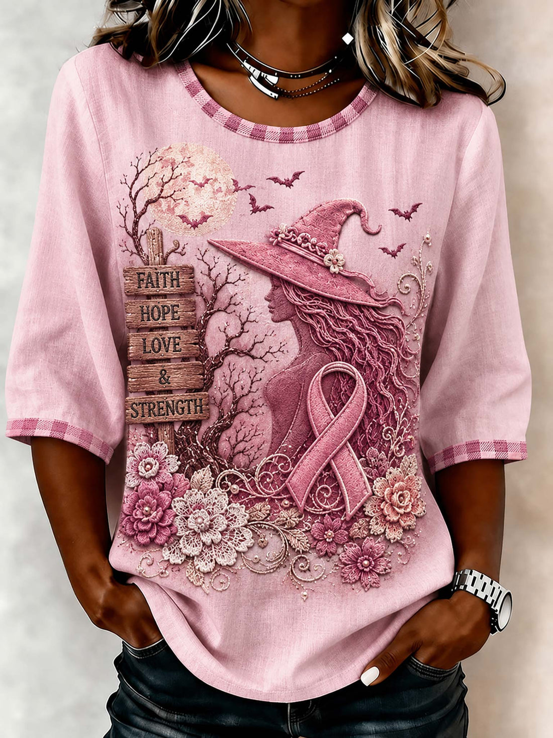 A woman wearing a pink 3/4 sleeve graphic t-shirt with a witch, pink ribbon, flowers, and the words 'FAITH HOPE LOVE & STRENGTH' printed on the front.