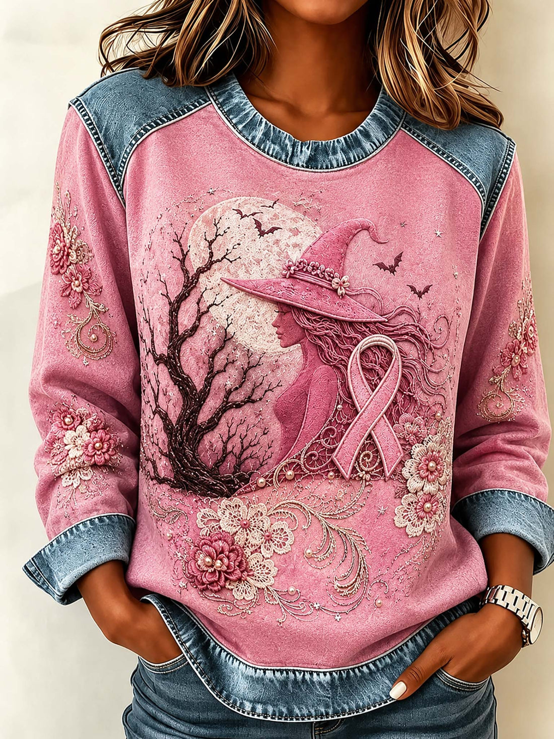 A woman wearing a pink long-sleeve sweatshirt with denim-style shoulder panels and cuffs, featuring a large graphic of a witch in a pink hat and pink ribbon under a tree, paired with blue jeans.