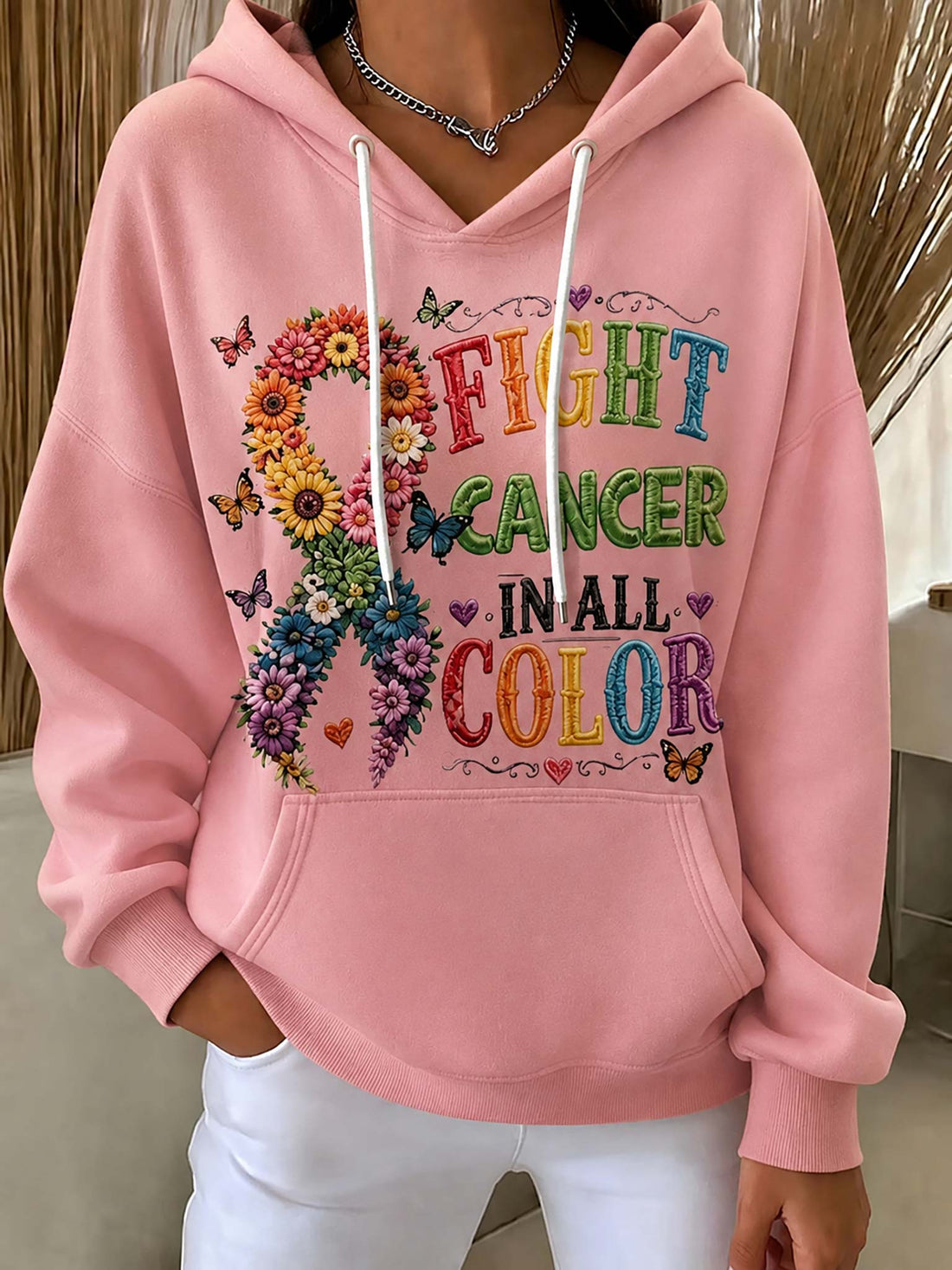 A person wearing a pink pullover hoodie with a large colorful graphic on the front that reads “FIGHT CANCER IN ALL COLOR” alongside a flower-and-butterfly ribbon design, paired with white pants.