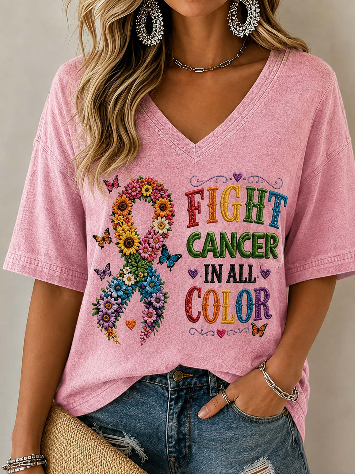 A woman wearing a pink V-neck t-shirt with a colorful graphic that reads 'FIGHT CANCER IN ALL COLOR' alongside a flower-and-butterfly awareness ribbon, paired with blue jeans.

