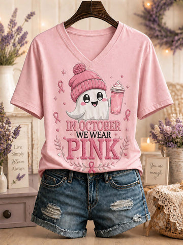 A pink V-neck graphic T-shirt displayed on a hanger with blue denim shorts. The shirt features a cartoon ghost wearing a pink beanie and holding a pink drink, with the text 'IN OCTOBER WE WEAR PINK' and pink ribbon motifs.