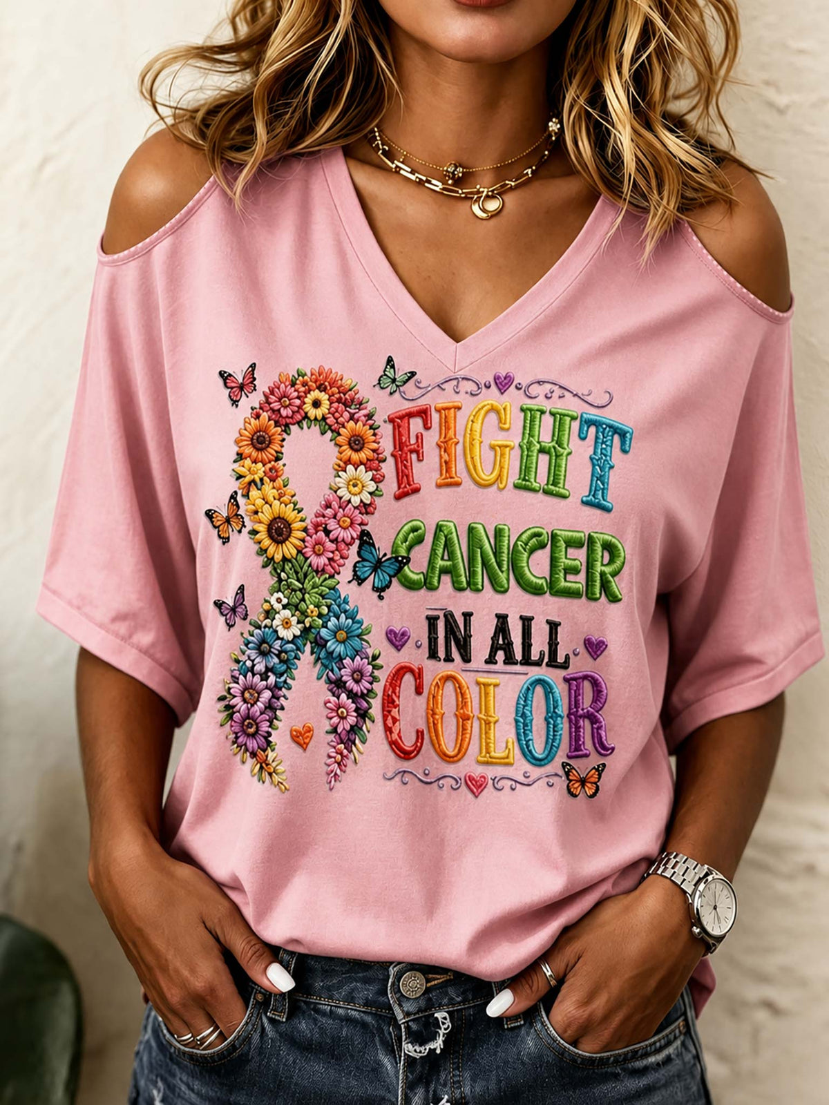 A woman wearing a pink cold-shoulder V-neck T-shirt with a colorful 'FIGHT CANCER IN ALL COLOR' graphic featuring a flower ribbon and butterflies, paired with blue jeans.