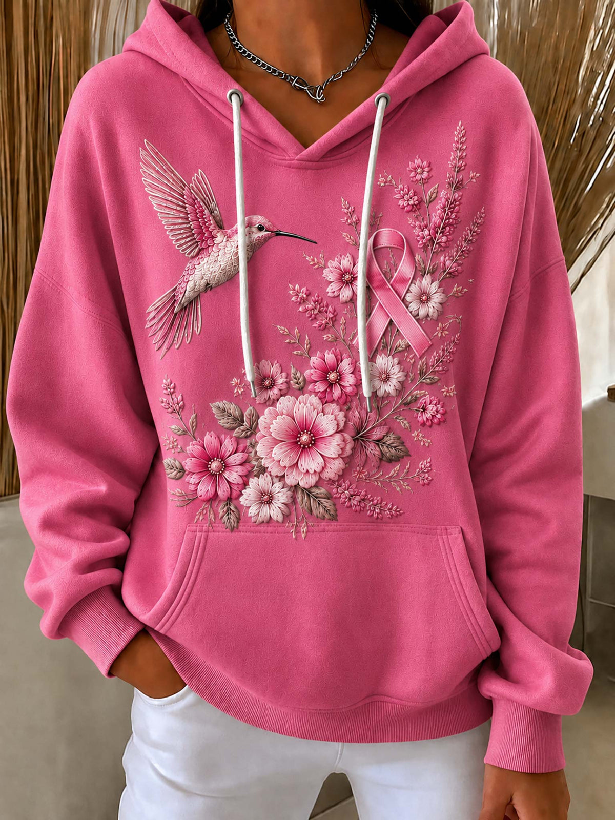 A pink pullover hoodie with a front graphic featuring a hummingbird, pink ribbon, and floral embroidery, worn by a model in a neutral indoor setting.