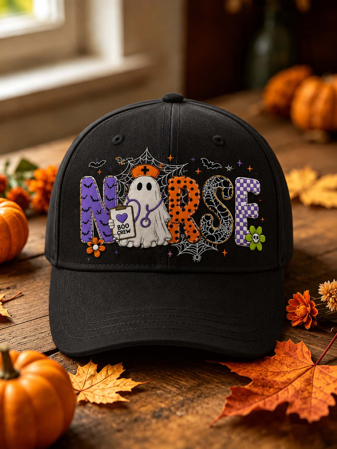 A black baseball cap with a Halloween-themed 'Nurse' graphic, displayed on a rustic wooden table surrounded by autumn leaves, pumpkins, and a window in the background.