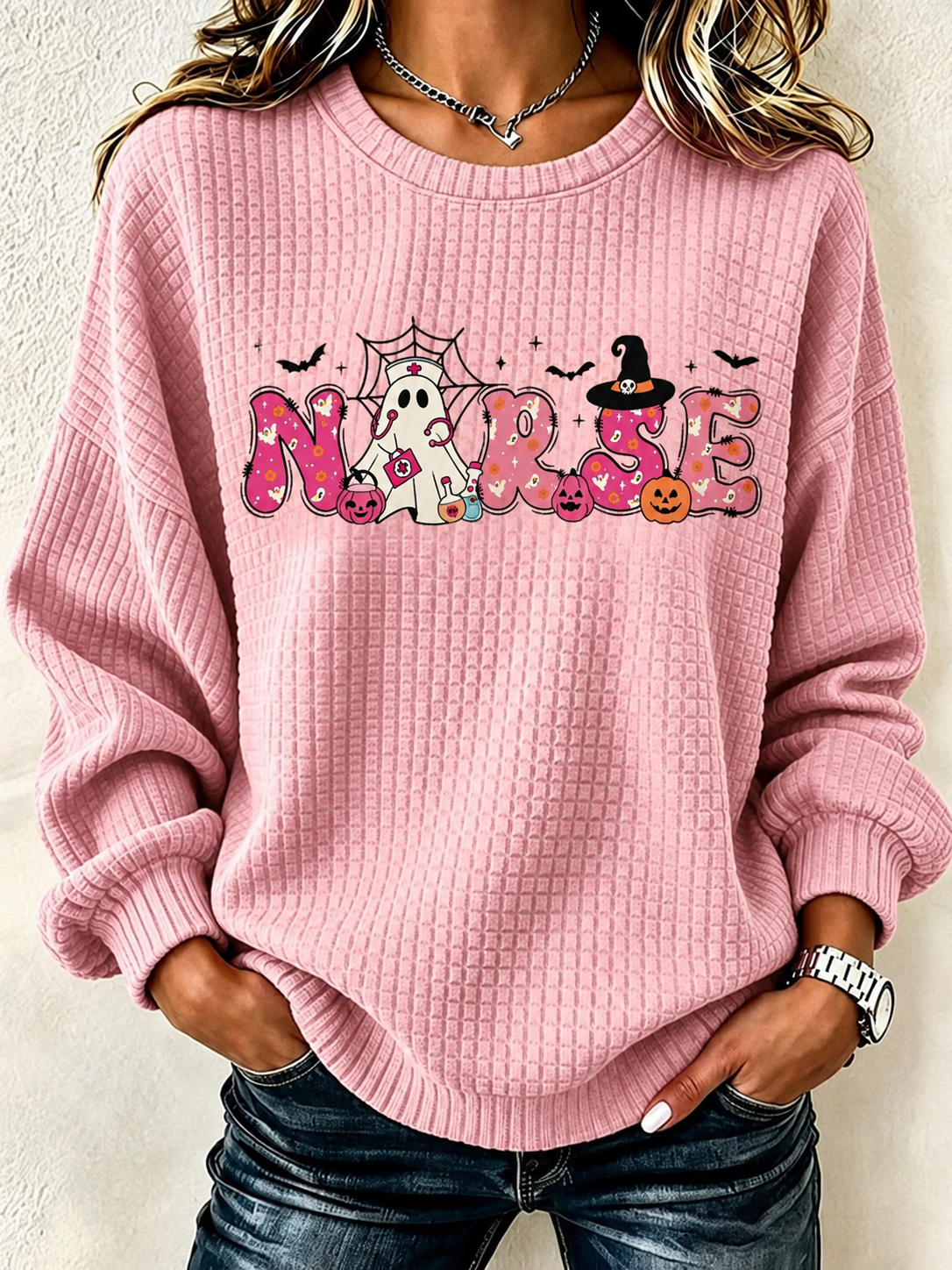 A woman wearing a pink waffle-knit crewneck sweatshirt with a 'Nurse' Halloween graphic, paired with dark blue jeans.