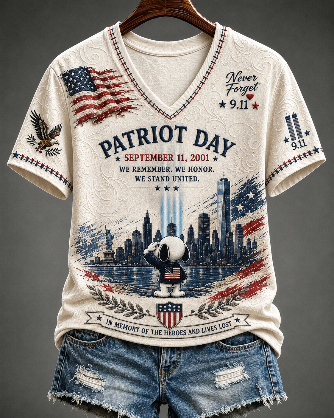 A cream-colored V-neck graphic t-shirt featuring a patriotic 'Patriot Day' design with Snoopy and a saluting dog, set against a gray studio background. The shirt is styled with distressed denim shorts on a wooden hanger.