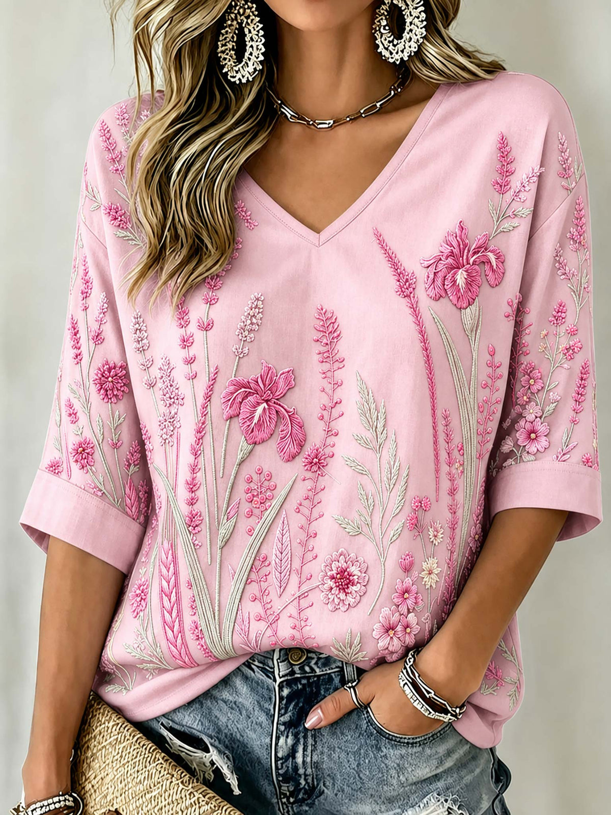 A woman wearing a light pink V-neck top with detailed pink and green floral embroidery, paired with distressed blue jeans and silver jewelry.