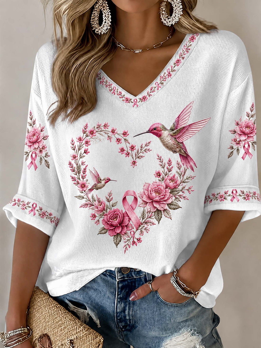 A woman wearing a white waffle-knit V-neck top with pink floral and hummingbird embroidery, paired with distressed blue jeans.