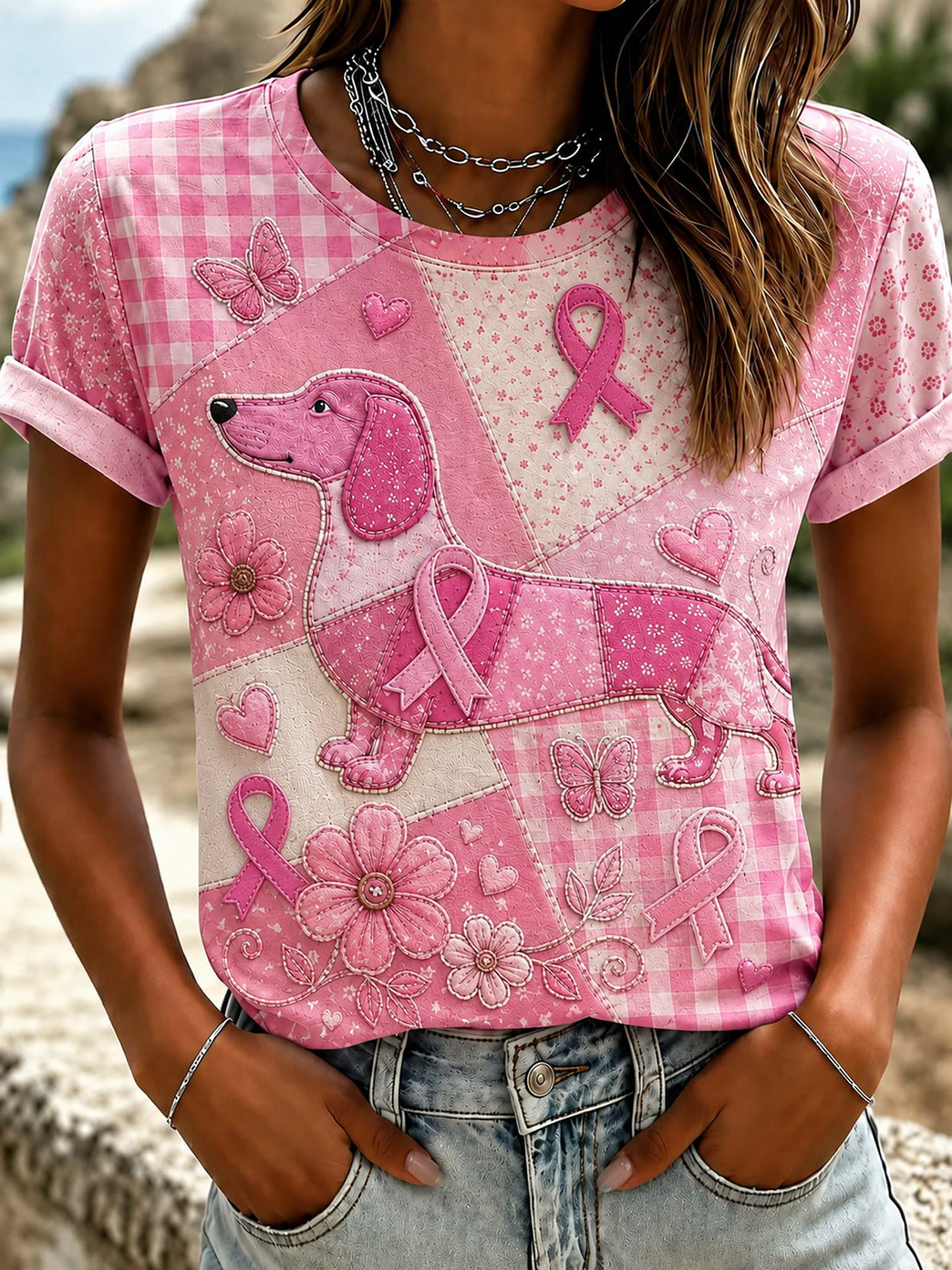 A pink short-sleeve t-shirt with a patchwork design featuring a dachshund, pink ribbons, flowers, butterflies, and gingham patterns, worn by a model in front of a blurred outdoor background.