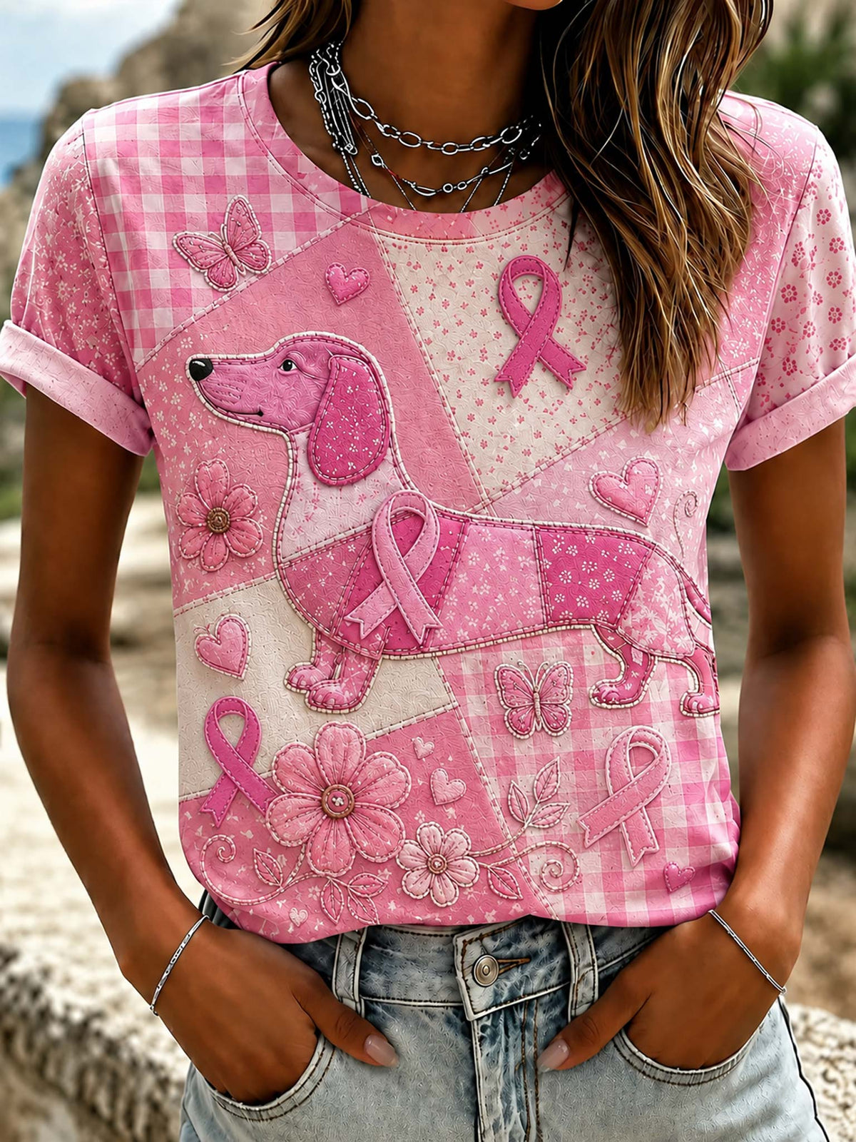 A pink short-sleeve t-shirt with a patchwork design featuring a dachshund, pink ribbons, flowers, butterflies, and gingham patterns, worn by a model in front of a blurred outdoor background.