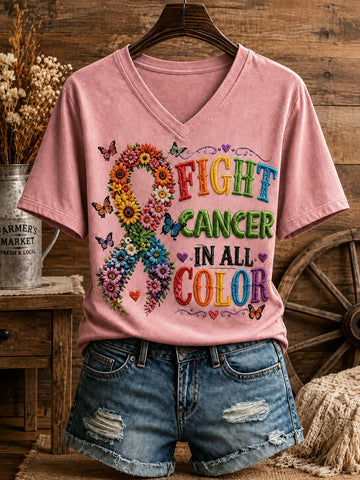 A pink V-neck graphic T-shirt displayed on a hanger with distressed denim shorts, set against a rustic wood-and-farm-themed backdrop. The shirt features a colorful floral awareness ribbon and the text 'FIGHT CANCER IN ALL COLOR' in multicolored lettering.
