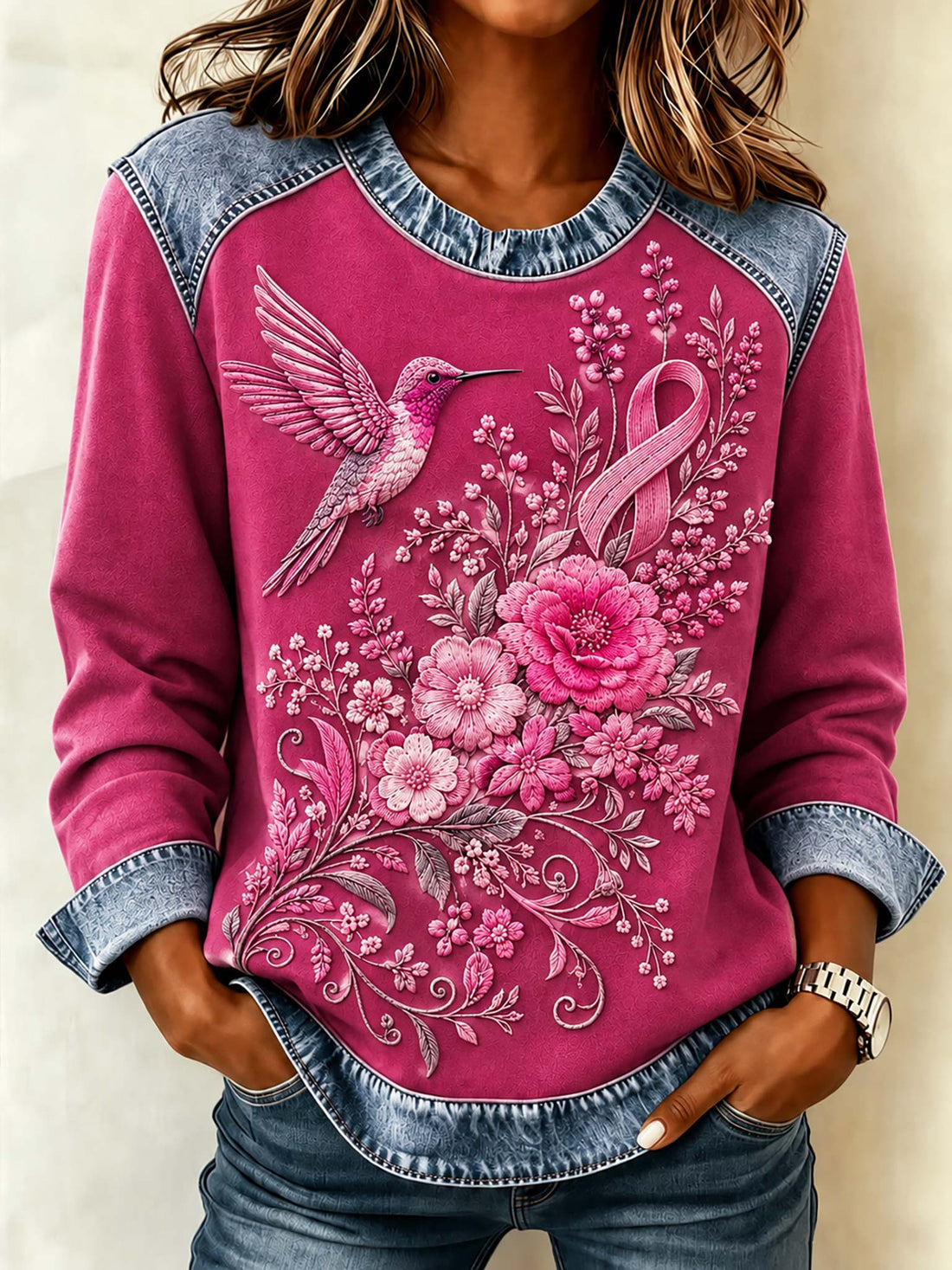 A woman wearing a long-sleeve crewneck sweatshirt with a vibrant pink body, denim blue sleeves and hem, and a detailed graphic of a hummingbird, flowers, and a pink awareness ribbon.