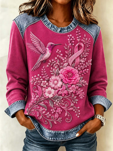 A woman wearing a long-sleeve crewneck sweatshirt with a vibrant pink body, denim blue sleeves and hem, and a detailed graphic of a hummingbird, flowers, and a pink awareness ribbon.