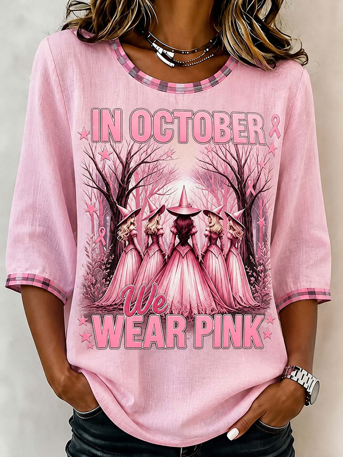 A woman wearing a light pink short-sleeve t-shirt with a graphic print that reads 'IN OCTOBER WE WEAR PINK' and shows three witches in pink robes standing in a forest. The t-shirt has plaid trim on the collar and cuffs.
