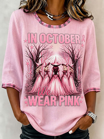 A woman wearing a light pink short-sleeve t-shirt with a graphic print that reads 'IN OCTOBER WE WEAR PINK' and shows three witches in pink robes standing in a forest. The t-shirt has plaid trim on the collar and cuffs.