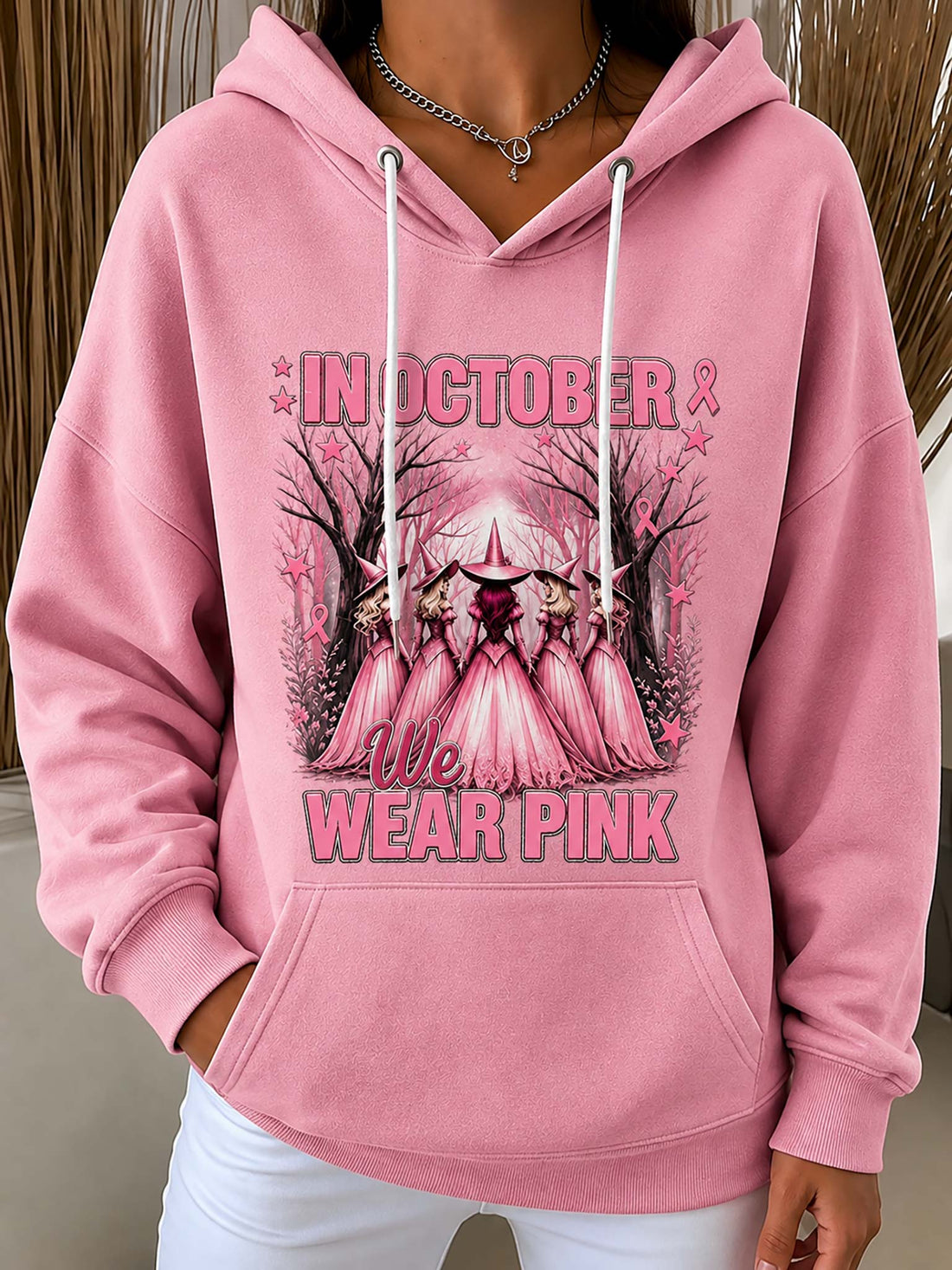 A pink pullover hoodie with a graphic print on the front that reads 'IN OCTOBER WE WEAR PINK' with a stylized pink cathedral and rose details, worn by a model.