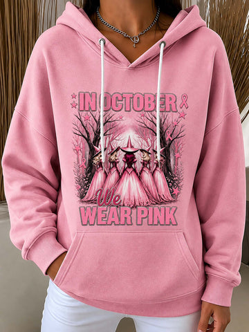 A pink pullover hoodie with a graphic print on the front that reads 'IN OCTOBER WE WEAR PINK' with a stylized pink cathedral and rose details, worn by a model.