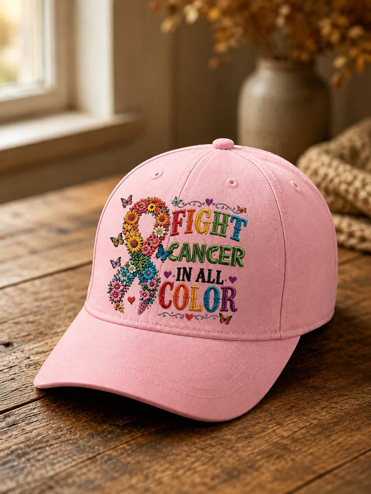A pink baseball cap resting on a rustic wooden table, featuring a colorful embroidered design with a floral ribbon and the words 'FIGHT CANCER IN ALL COLOR'.