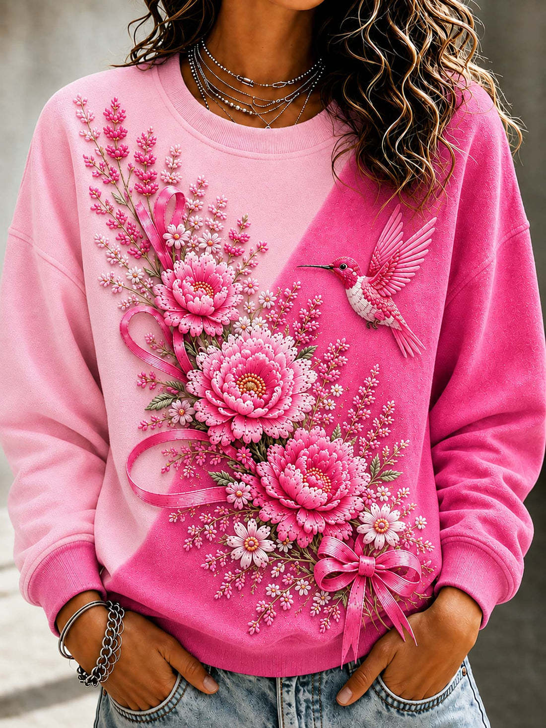 A close-up of a person wearing a pink crewneck sweatshirt with a large 3D floral and hummingbird applique design on the front, paired with blue jeans.