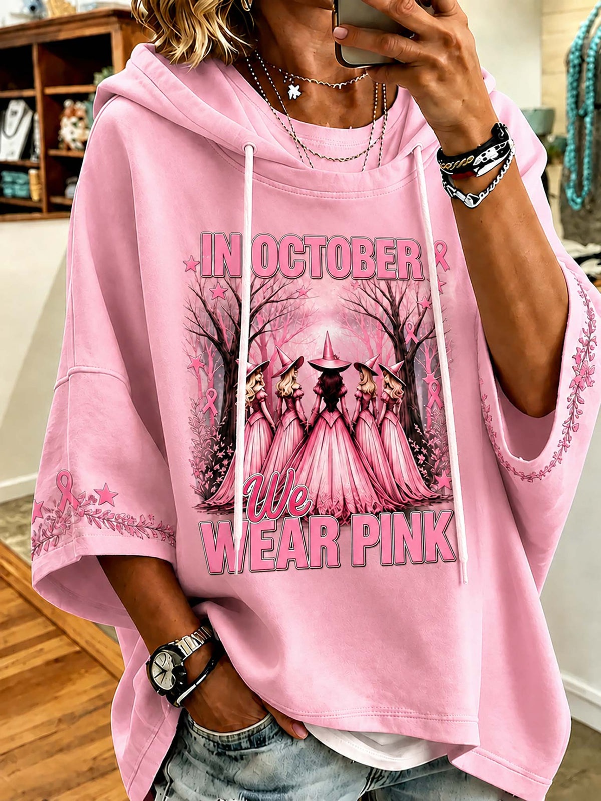 A person wearing a pink oversized hoodie with a graphic print that reads 'IN OCTOBER WE AR PINK' and shows a mirror selfie in an indoor setting.