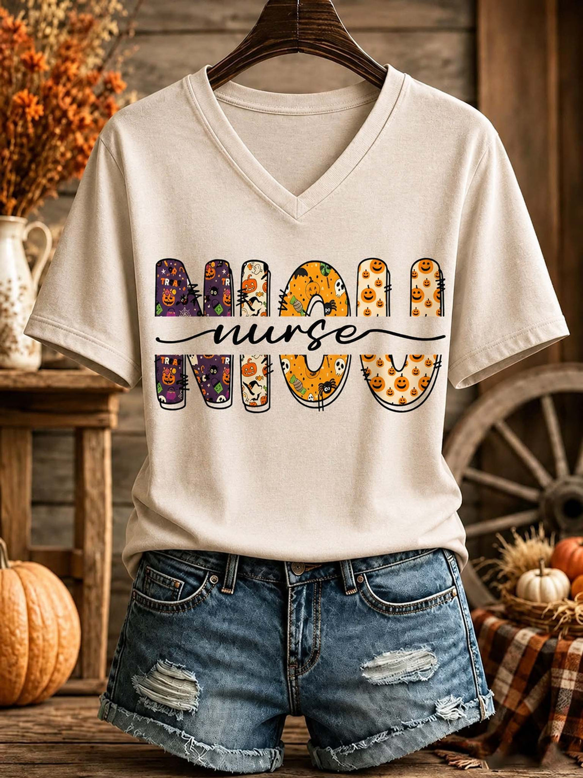 A beige V-neck t-shirt with a 'MOM NURSE' graphic featuring Halloween-themed letter designs, styled on a hanger with denim shorts against a rustic fall-themed background.