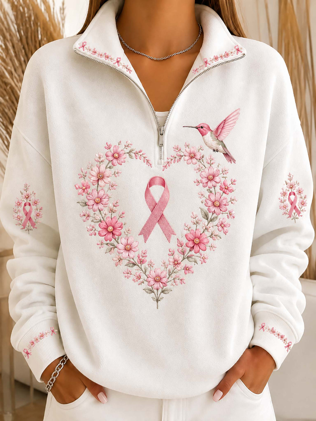 A woman wearing a white half-zip sweatshirt with a pink ribbon and floral heart design on the front, along with a pink hummingbird graphic. The sweatshirt features pink floral embroidery details on the collar, cuffs, and sleeves.