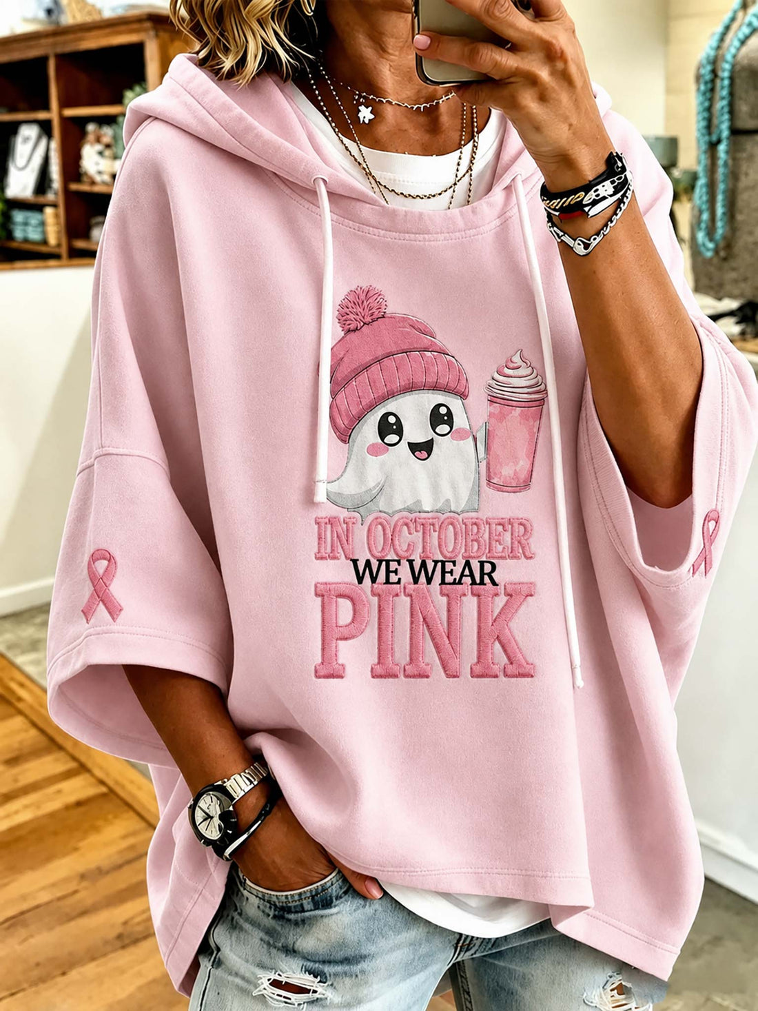 A woman wearing a light pink oversized hoodie with a cute cartoon ghost wearing a pink beanie and holding a pink drink. The hoodie features the text 'IN OCTOBER WE WEAR PINK' on the front, with a pink ribbon printed on the sleeve.