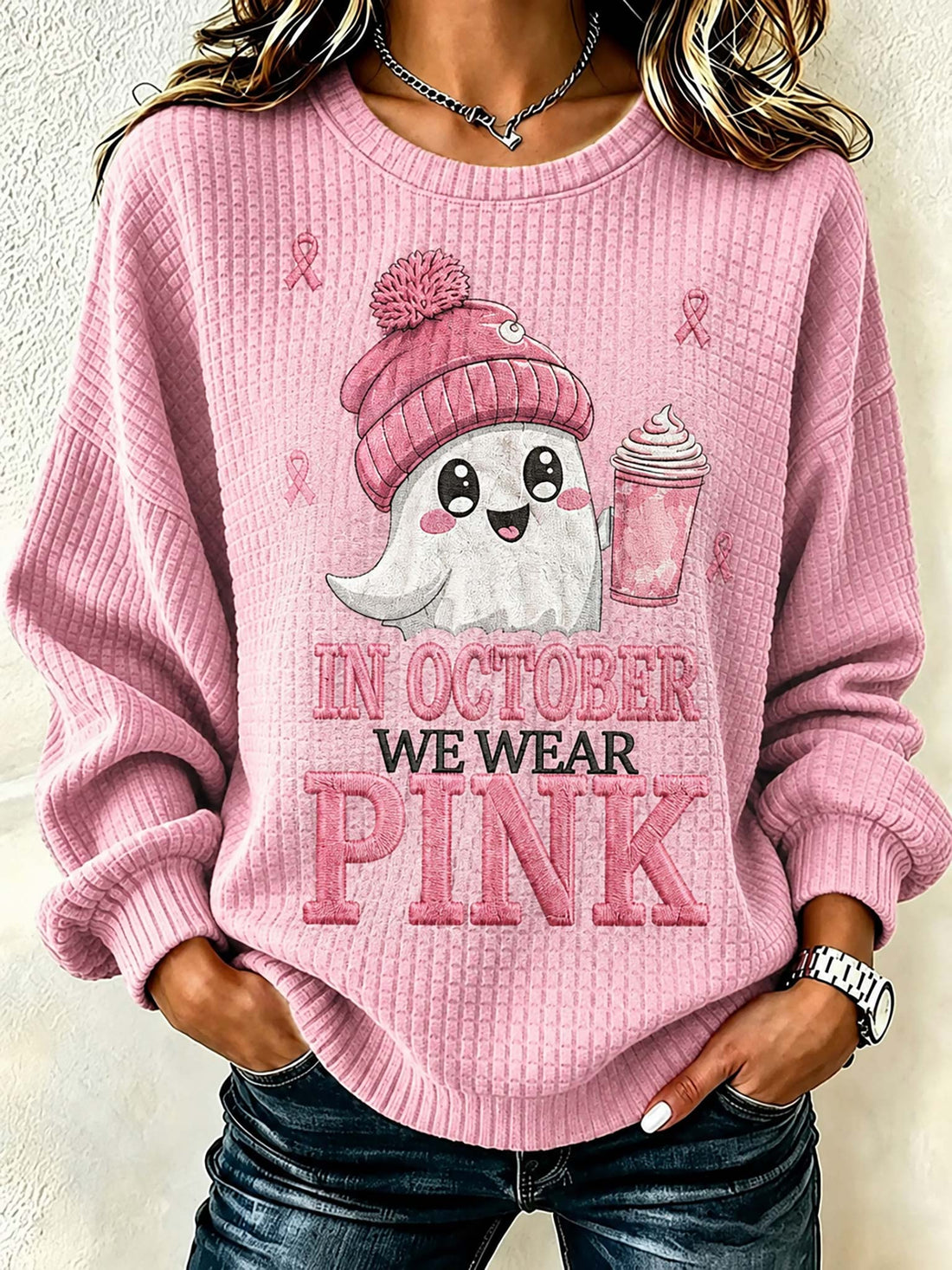 A woman wearing a pink waffle-knit crewneck sweatshirt with a graphic of a cute ghost in a pink beanie holding a pink drink, with the text 'IN OCTOBER WE WEAR PINK' printed on the front.