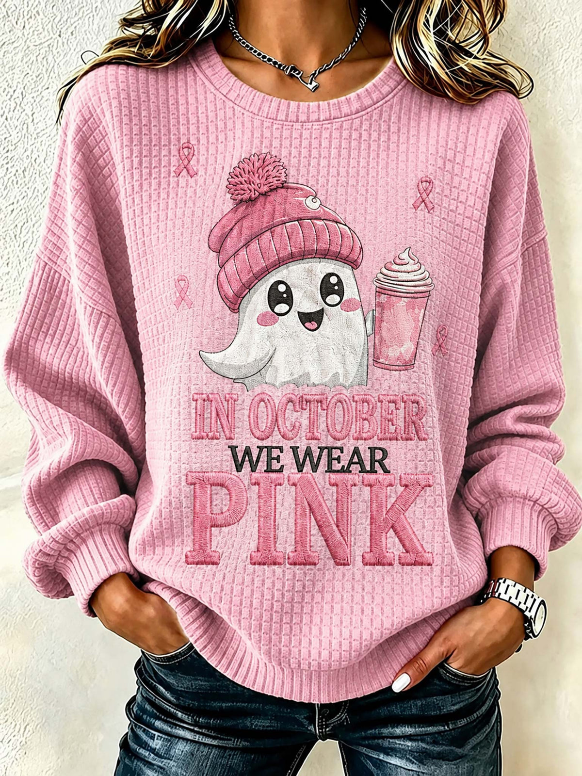 A woman wearing a pink waffle-knit crewneck sweatshirt with a graphic of a cute ghost in a pink beanie holding a pink drink, with the text 'IN OCTOBER WE WEAR PINK' printed on the front.