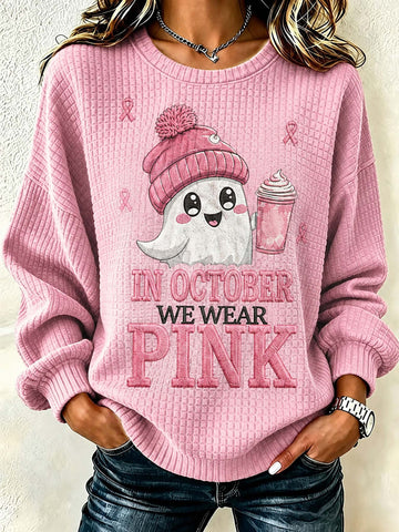 A woman wearing a pink waffle-knit crewneck sweatshirt with a graphic of a cute ghost in a pink beanie holding a pink drink, with the text 'IN OCTOBER WE WEAR PINK' printed on the front.