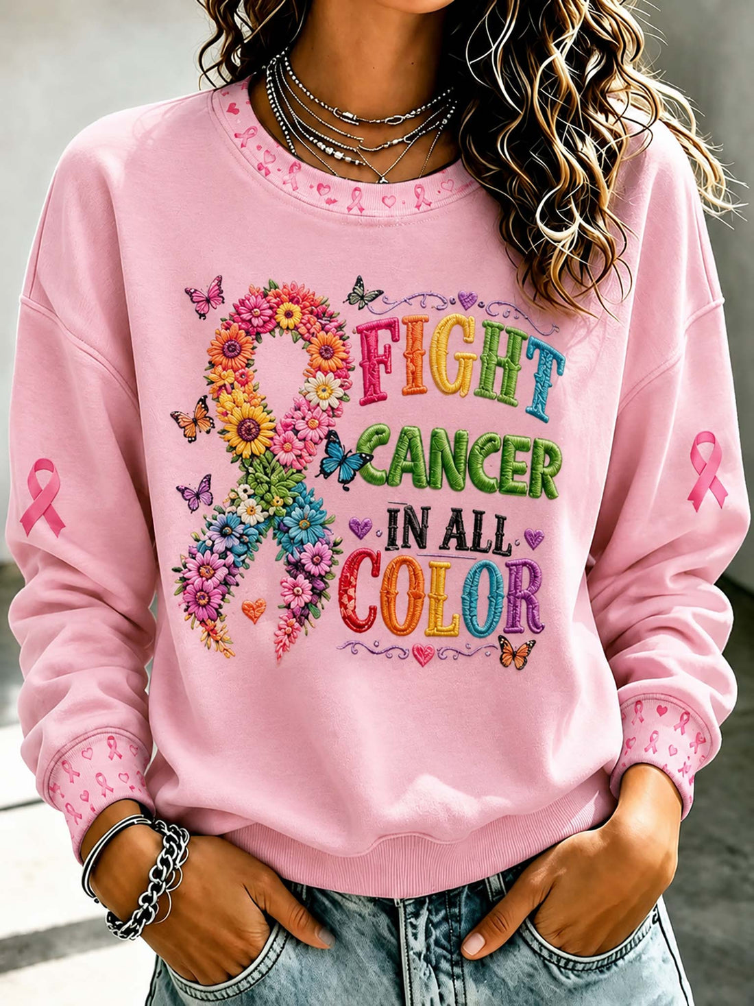 A woman wearing a light pink crewneck sweatshirt with a colorful graphic that reads 'FIGHT CANCER IN ALL COLOR' alongside a flower and ribbon design, paired with light blue jeans.

