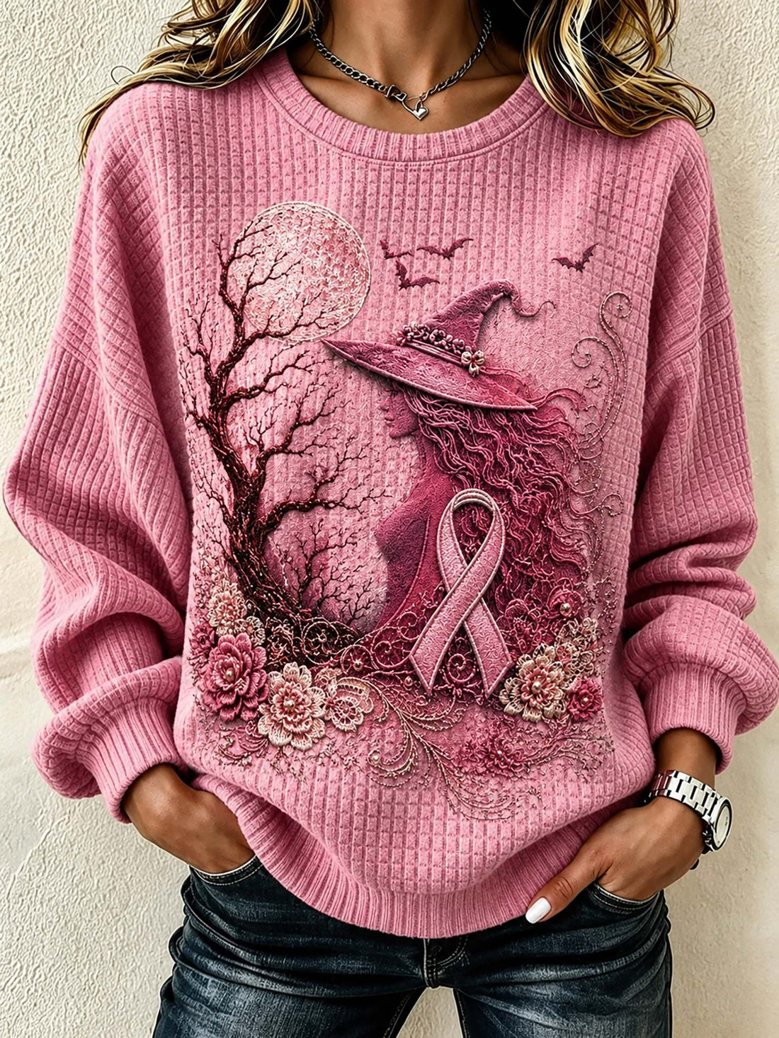 A woman wearing a pink ribbons-of-dignity themed sweater with a witch, tree, and rose graphic, paired with dark blue jeans.