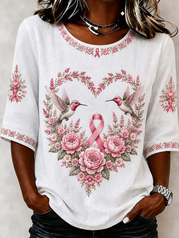 A woman wearing a white short-sleeve t-shirt with a pink breast cancer awareness graphic featuring two hummingbirds, pink roses, and a central pink ribbon, framed by a floral heart design.
