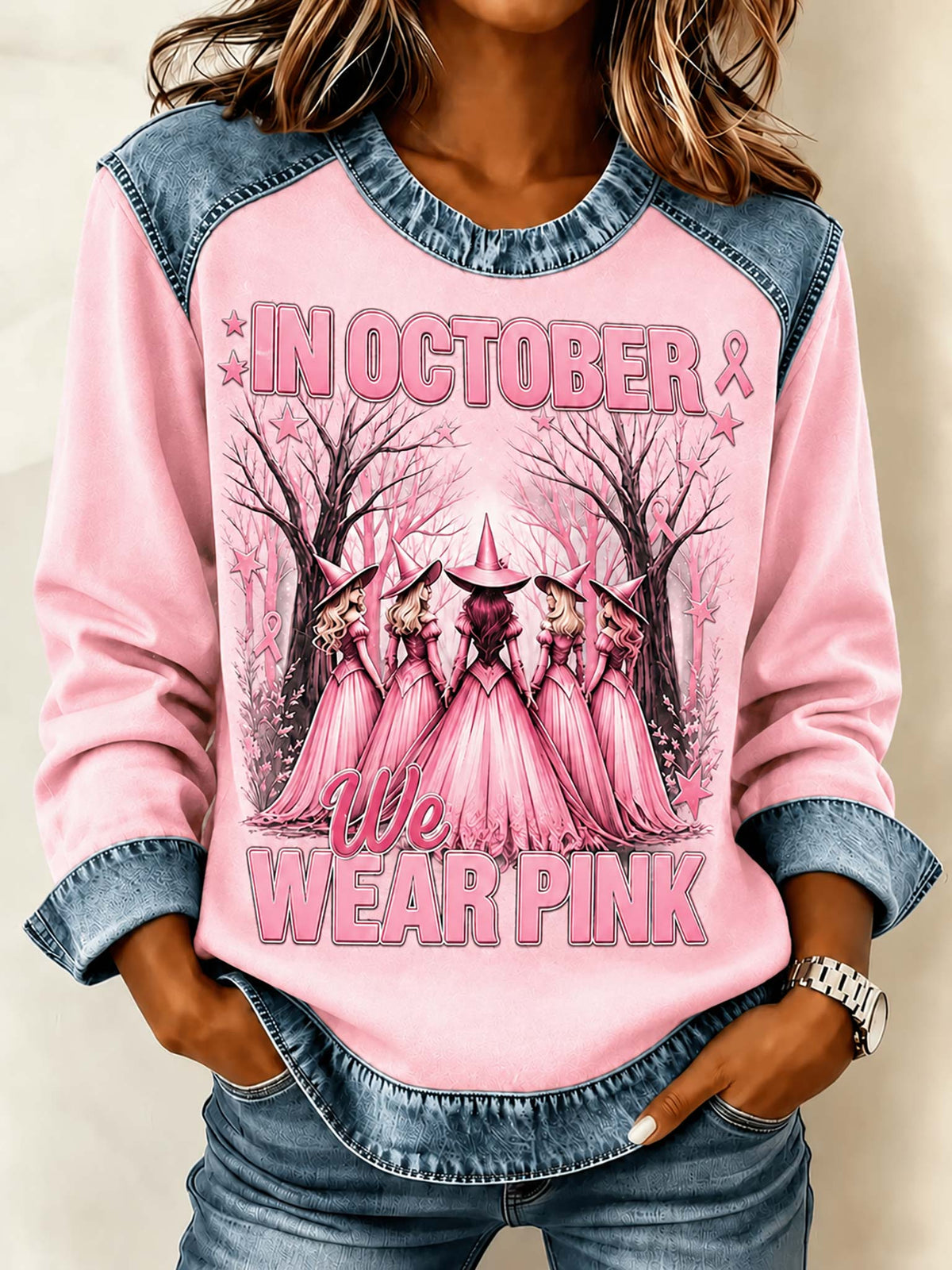 A woman wearing a pink raglan long-sleeve shirt with denim shoulder panels and cuffs, featuring a graphic of pink-cloaked figures in a forest with the text 'IN OCTOBER WE WEAR PINK'.