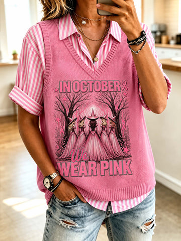 A woman takes a mirror selfie wearing a pink knit V-neck sweater vest over a pink and white striped button-down shirt, paired with distressed blue jeans. The vest features a graphic of three witches in pink robes with the text 'IN OCTOBER We WEAR PINK'.