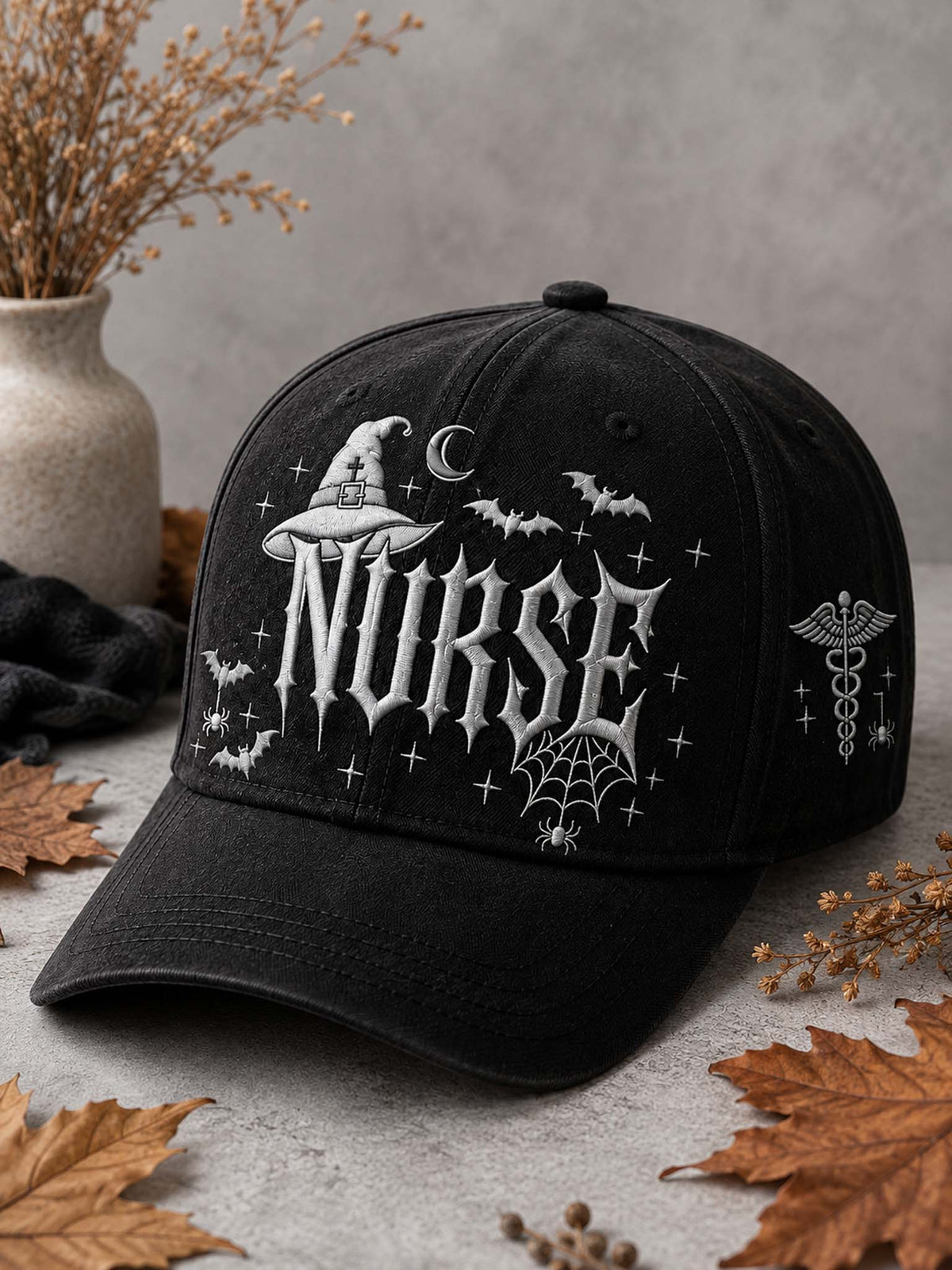 A black baseball cap with gothic Halloween-themed embroidery, featuring the word 'NURSE' in a stylized font with a witch hat, bats, spiderwebs, and a caduceus symbol. The cap is styled on a gray surface with autumn leaves and a vase of dried grass in the background.