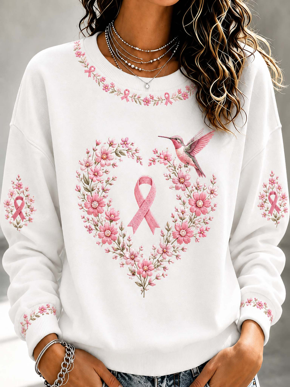 A woman wearing a white crewneck sweatshirt with a pink breast cancer awareness design featuring a pink ribbon, a floral heart, and a hummingbird.
