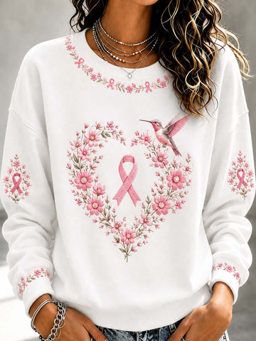 A woman wearing a white crewneck sweatshirt with a pink breast cancer awareness design featuring a pink ribbon, a floral heart, and a hummingbird.