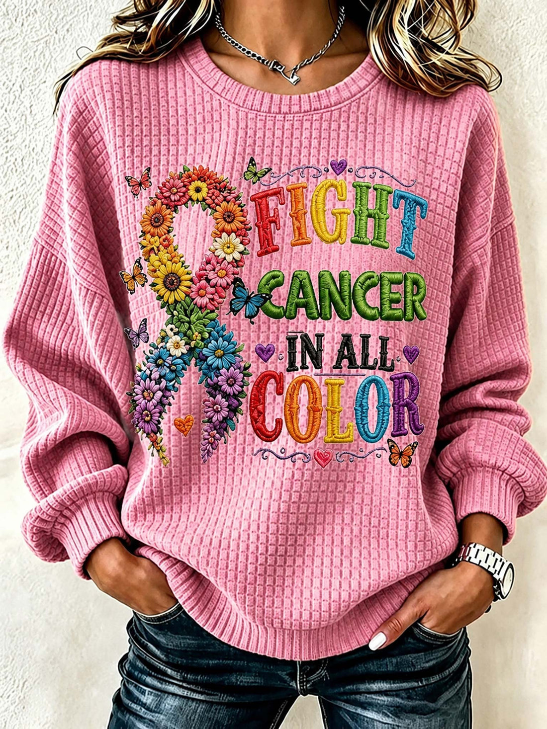 A woman wearing a pink waffle-knit crewneck sweatshirt with a colorful graphic that reads 'FIGHT CANCER IN ALL COLOR' alongside a flower-and-butterfly awareness ribbon, paired with dark blue jeans.
