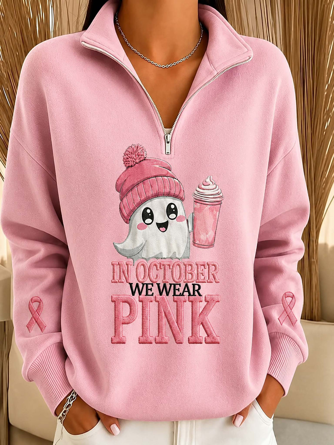 A person wearing a pink quarter-zip sweatshirt with a graphic of a ghost in a pink beanie holding a pink drink, with the text 'IN OCTOBER WE WEAR PINK' on the front. Pink awareness ribbons are printed on both sleeves.