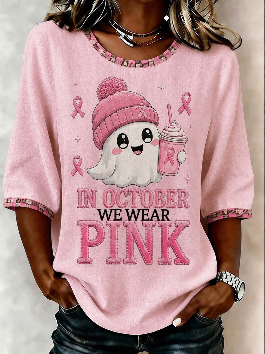 A woman wearing a pink short-sleeve graphic T-shirt with a cartoon ghost wearing a pink beanie and holding a pink drink, with the text 'IN OCTOBER WE WEAR PINK' on the front.