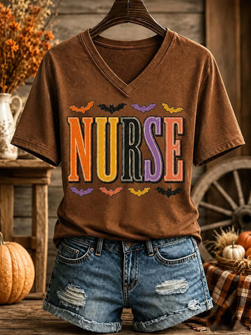 A brown V-neck graphic T-shirt on a hanger, styled with distressed denim shorts in a rustic fall-themed setting. The shirt features the word 'NURSE' in large, colorful block letters with small bat silhouettes above and below.