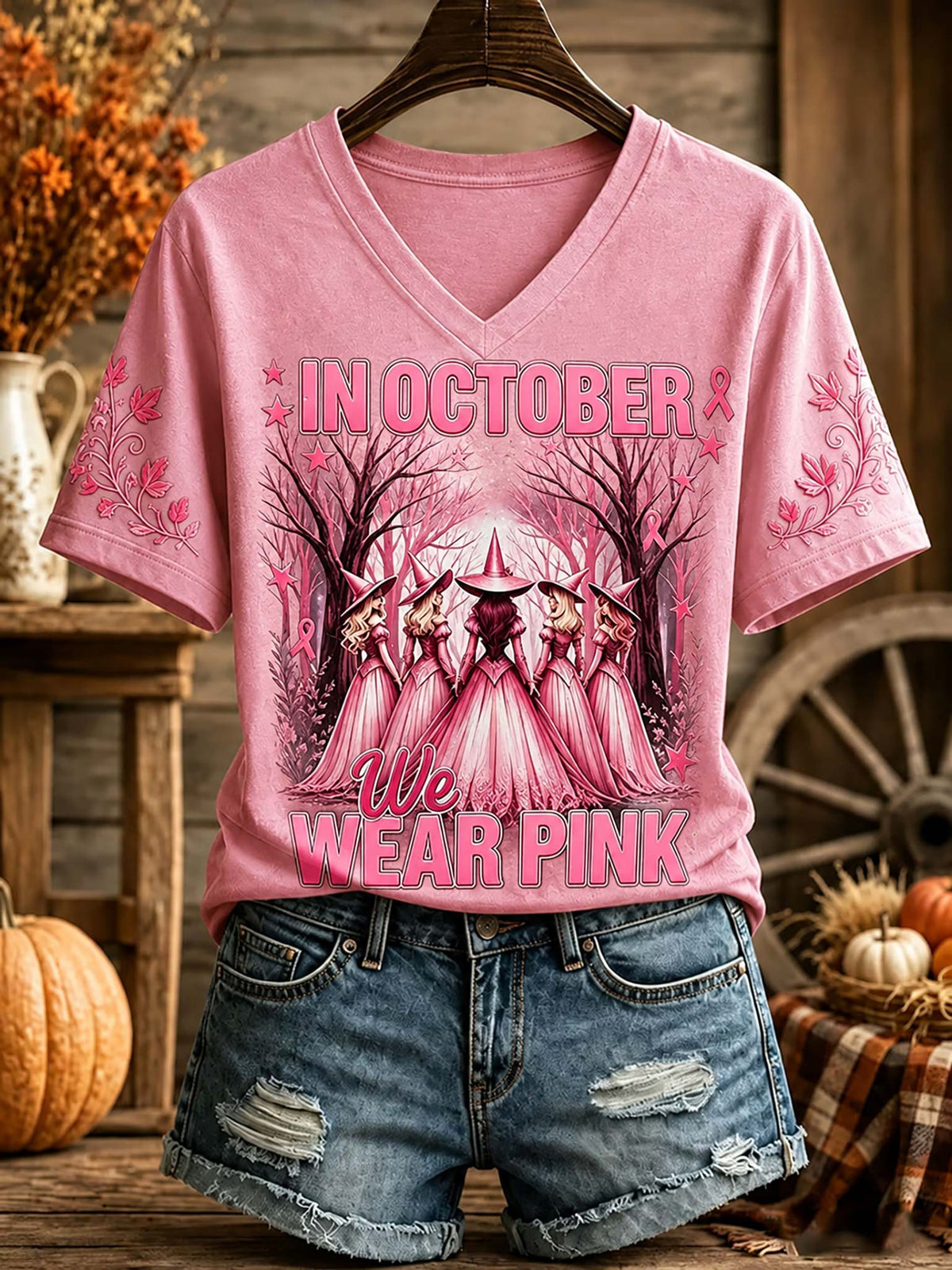 A pink V-neck graphic T-shirt displayed on a hanger with blue denim shorts, set against a rustic autumn-themed background with pumpkins, a wagon wheel, and fall decor. The shirt features a graphic of witches in pink hats and cloaks with the text 'IN OCTOBER We WEAR PINK' and pink ribbon motifs on the sleeves.