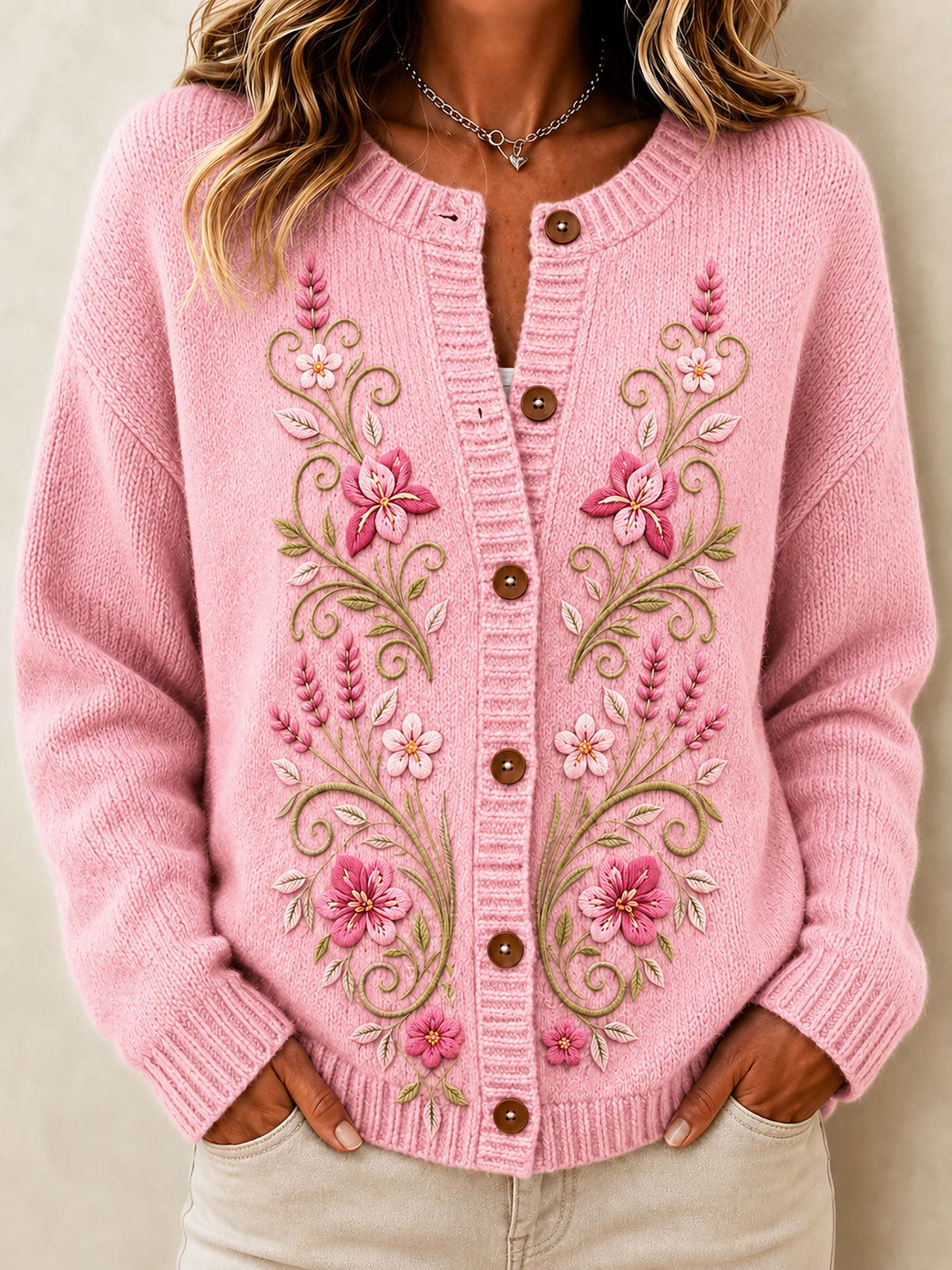 A woman wearing a pink knit cardigan sweater with floral embroidery and brown buttons, paired with light beige pants, against a neutral beige background.