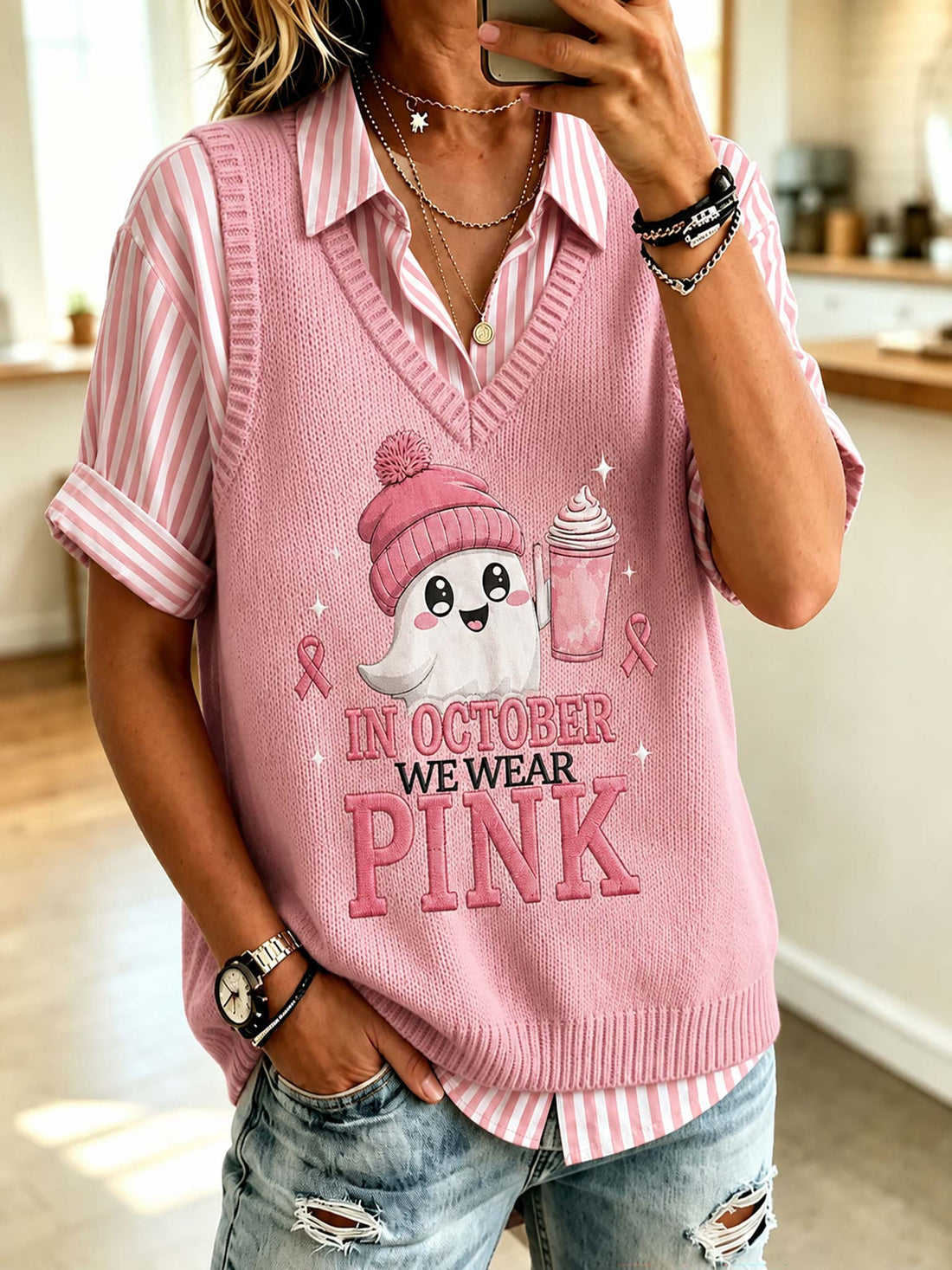 A woman wearing a pink knit sweater vest over a pink and white striped short-sleeve collared shirt, paired with distressed blue jeans. The vest features a cartoon ghost wearing a pink beanie and the text 'IN OCTOBER WE WEAR PINK'.