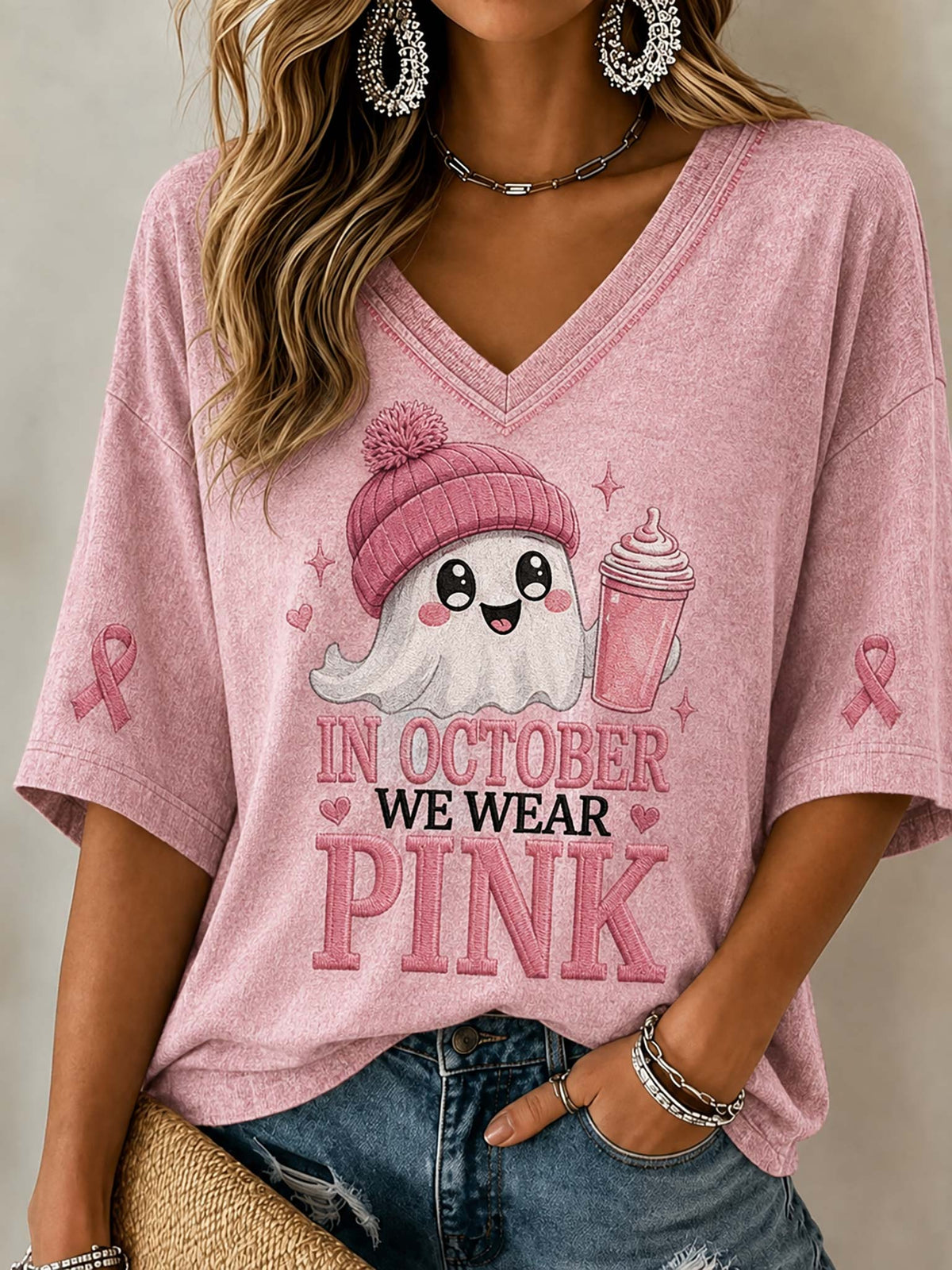 A woman wearing a pink V-neck short-sleeve t-shirt with a cartoon ghost graphic and the text 'IN OCTOBER WE WEAR PINK' on the front, paired with blue jeans.