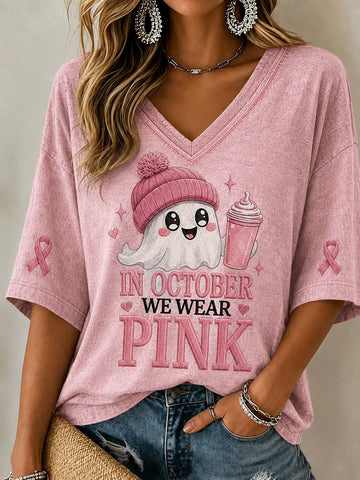 A woman wearing a pink V-neck short-sleeve t-shirt with a cartoon ghost graphic and the text 'IN OCTOBER WE WEAR PINK' on the front, paired with blue jeans.